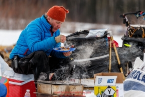 Linwood Fiedler sits outside at his sled and eats a homemade dinner of shrimp and quinoa he heated in his dog water at the Nikolai checkpoint during Iditarod 2016.  Alaska.  March 08, 2016.  Photo by Jeff Schultz (C) 2016 ALL RIGHTS RESERVED