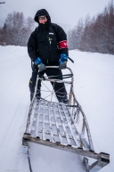 A Ruby veterinarian hitches a ride on a sled attached to a snow machine for a lift up the hill on March 14, 2020.
