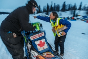 Volunteer checker Ann Ghicaduf delivers Heet and points Jason Campeau toward the water hole after Jason arrived in the morning at the Finger Lake checkpoint at Winterlake Lodge during Iditarod 2016.  Alaska.  March 07, 2016.  Photo by Jeff Schultz (C) 2016 ALL RIGHTS RESERVED